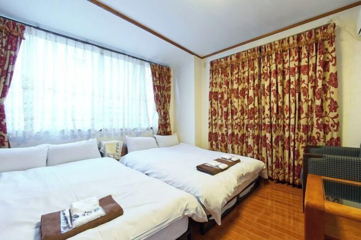 A hotel room with two beds, a wooden floor, and a view of the outdoors through sheer curtains.