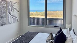 A bedroom with a large window, a bed with pillows, and artwork on the wall. - Sea Dunes Luxury Apartment - 10 steps to the beach (Moana)