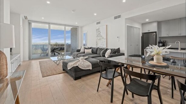 A modern living room with a sofa, dining table, and chairs, a kitchen area, and a balcony with a view. - Sea Dunes Luxury Apartment - 10 steps to the beach (Moana)