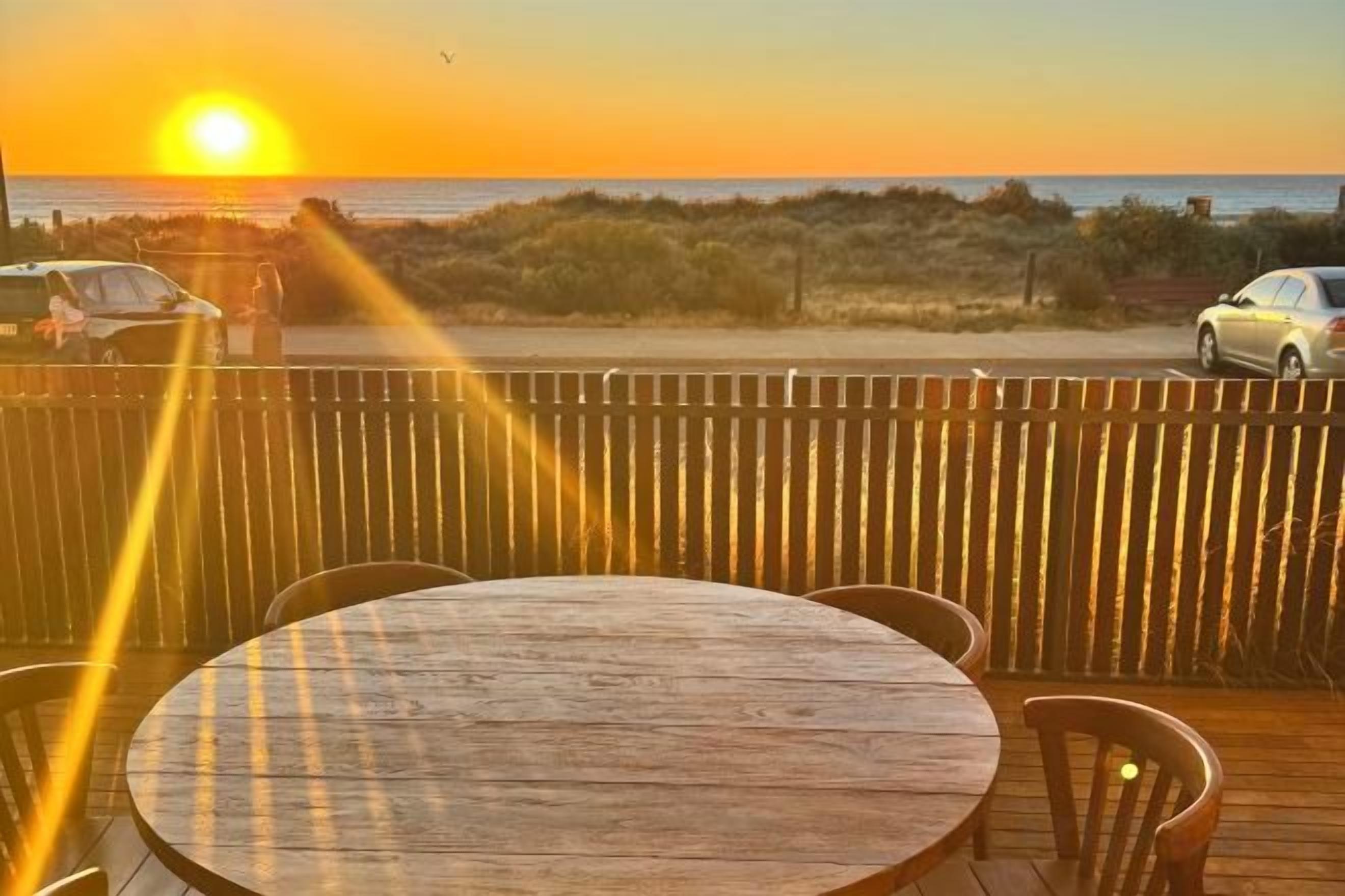 A wooden deck with a round table and chairs, overlooking a beach with a sunset.