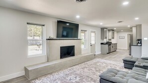 A spacious living room with a fireplace, a large flat-screen TV mounted above it, and a grey sectional sofa. - Backyard & Game Room! Home in Heart of Midland (Midland)