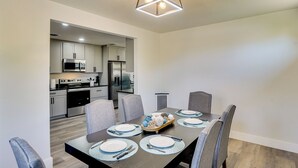 A modern dining area with a rectangular table set for six, surrounded by grey upholstered chairs. A kitchen is visible in the background with stainless steel appliances. - Backyard & Game Room! Home in Heart of Midland (Midland)