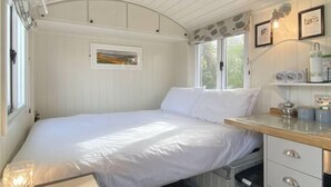 A single bed with white bedding, a wooden bedside table with a lamp, a framed picture on the wall, and a window with a view of greenery.