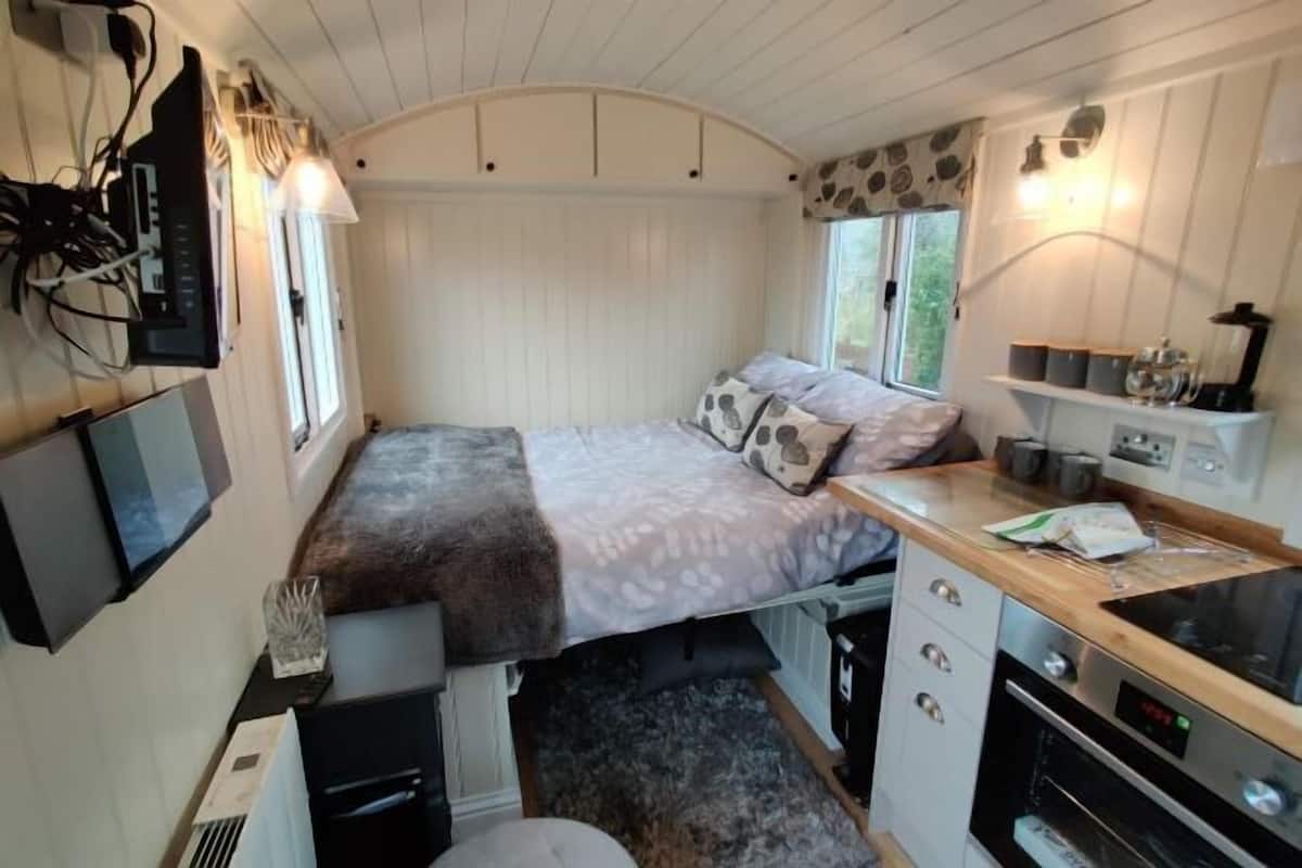 A compact RV interior with a bed, kitchenette, and a TV mounted on the wall.