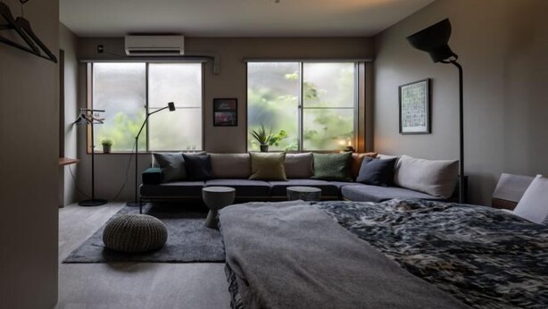 Una camera da letto moderna con un letto grande, un divano e un'area scrivania. Sono presenti due finestre con tende, una lampada da terra e un condizionatore d'aria a muro.