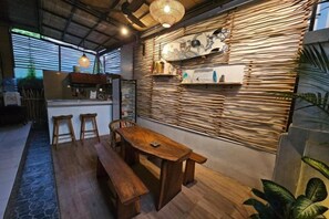 A dining area with a wooden bench and table, a wall-mounted shelf with decorative items, and a hanging light fixture. - Chris Homestay (Bali)