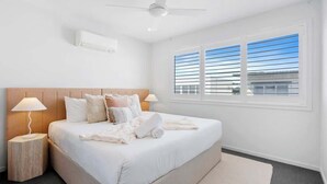 A hotel room with a bed, a ceiling fan, an air conditioning unit, a window with blinds, and a nightstand with a lamp. - Seabreeze (kingscliff)