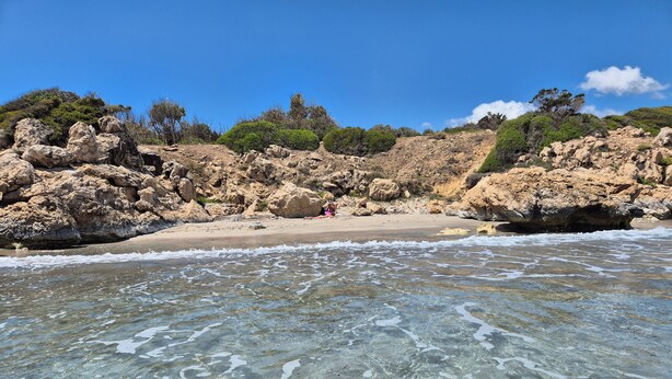 Una platja amb aigües poc profundes i cristal·lines, penya-segats rocosos i vegetació escassa.