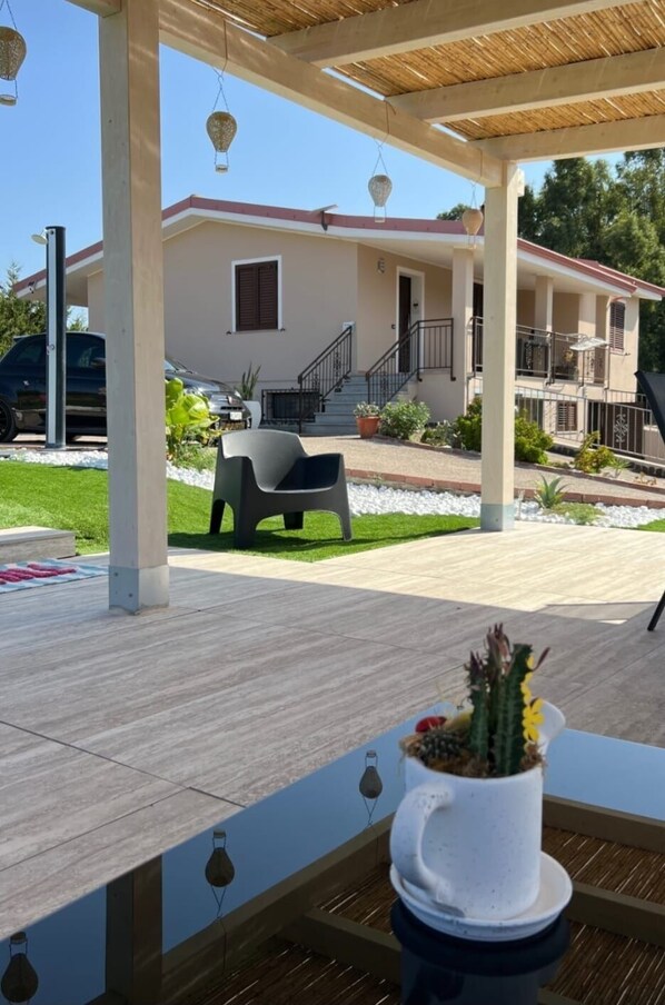 A covered outdoor area with a table, a potted plant, and a building in the background. - Family Villa in Southern Sardinia – Peace, Comfort, and Proximity to the Sea (Matzaccara)