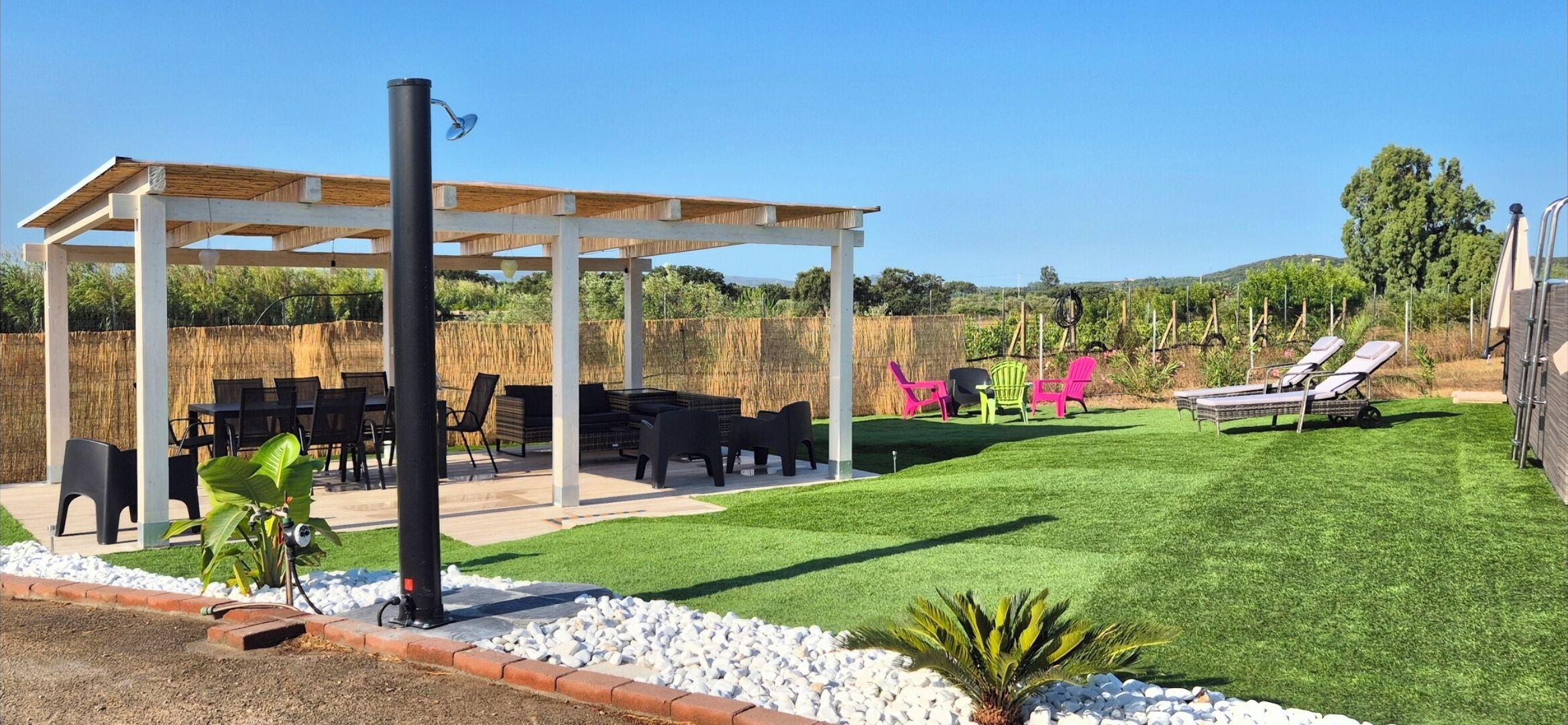 Uma área de estar coberta ao ar livre com mesas e cadeiras, um gramado e uma estrutura de pérgola.