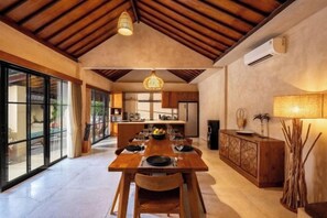 A spacious dining area with a wooden table set for a meal, a kitchen in the background, and a large window providing natural light. - Villa Terracotta by Pertama Stay (Bali)