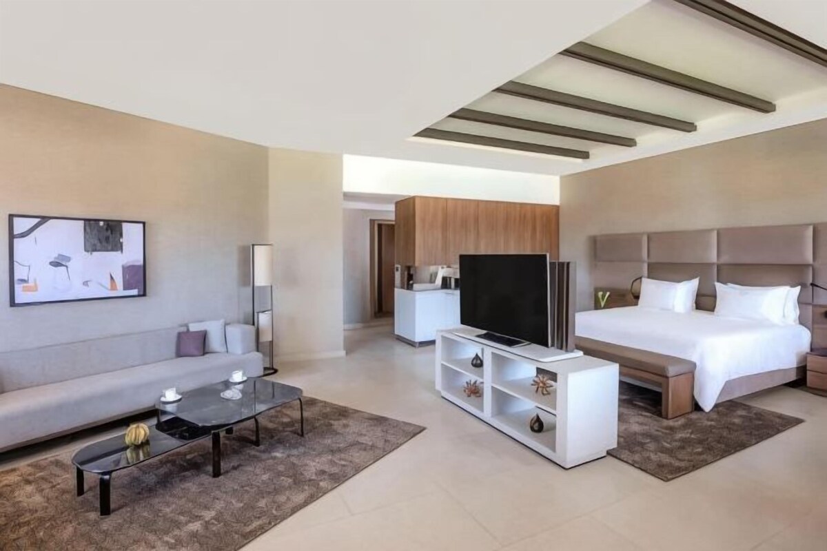 A modern hotel room with a bed, TV, sofa, and a dining area.