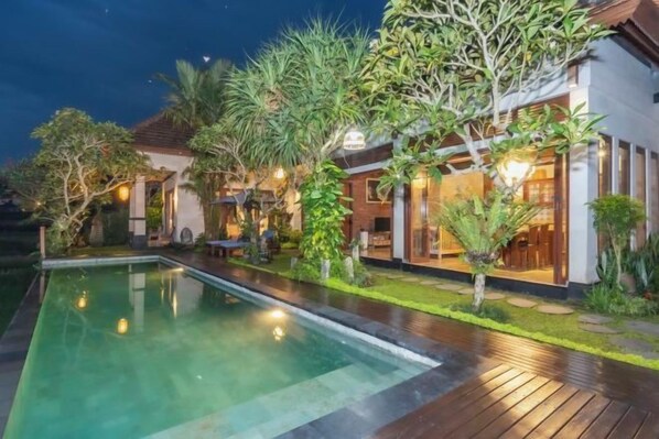 A pool area with a wooden deck, surrounded by tropical plants and a building with large windows. - Umah Sunset (Bali)