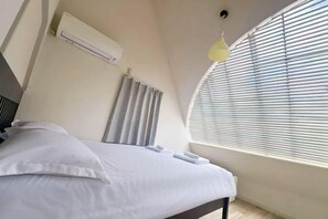 Ein Schlafzimmer mit einem Bett, weißer Bettwäsche, einem Deckenventilator, einer Klimaanlage, einem Fenster mit Jalousien und einer Leuchte.