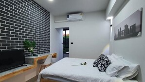 A bedroom with a brick-patterned wall, a bed with white bedding, a television on a wooden desk, and a painting on the wall. - Irich residence (South Samrong) 6 Kms BTS-Pu Chao (Phra Pradaeng)