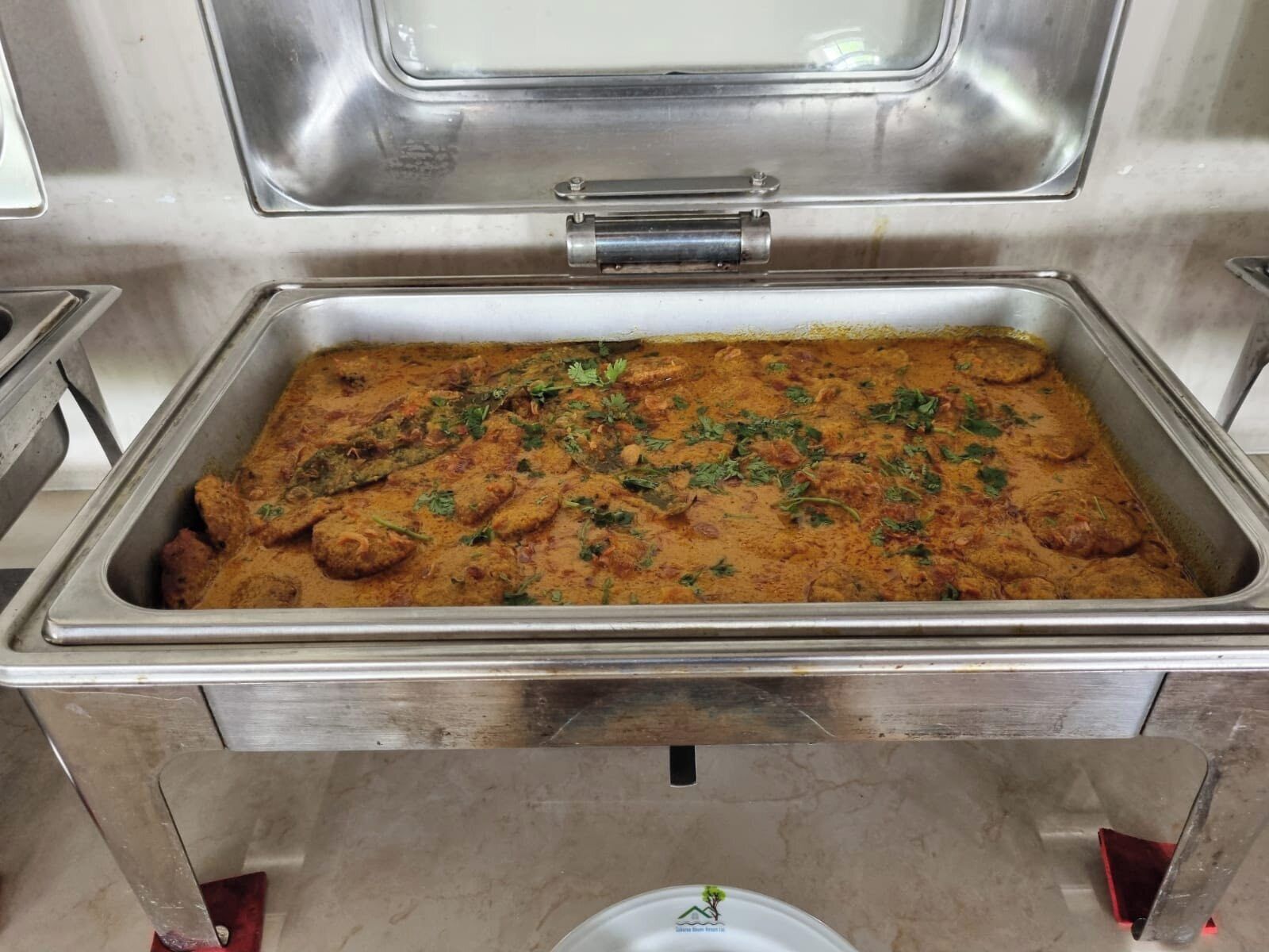 A buffet setup with a large metal serving tray filled with a curry dish, garnished with herbs.