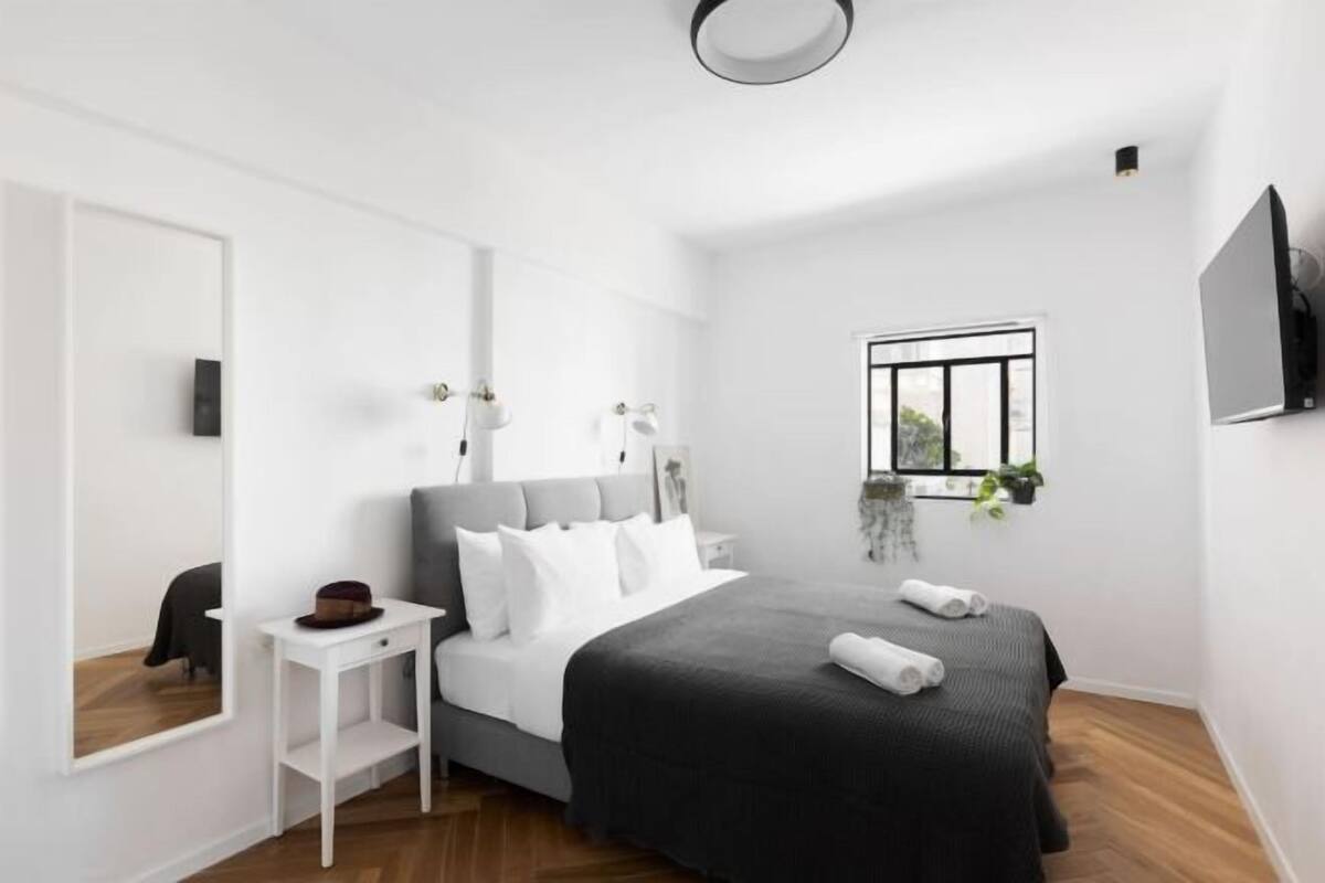 A modern bedroom with a bed, bedside table, and a window with plants.