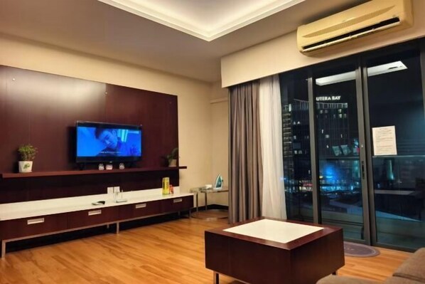 A modern living room with a flat-screen TV, a wooden entertainment unit, a glass door with a city view, and a small table. - South China Sea Place Suites by Kinabina Homestay (Kota Kinabalu)