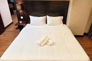A neatly made bed with white linens and a set of towels, a dark headboard, and a small bedside table with a lamp.
