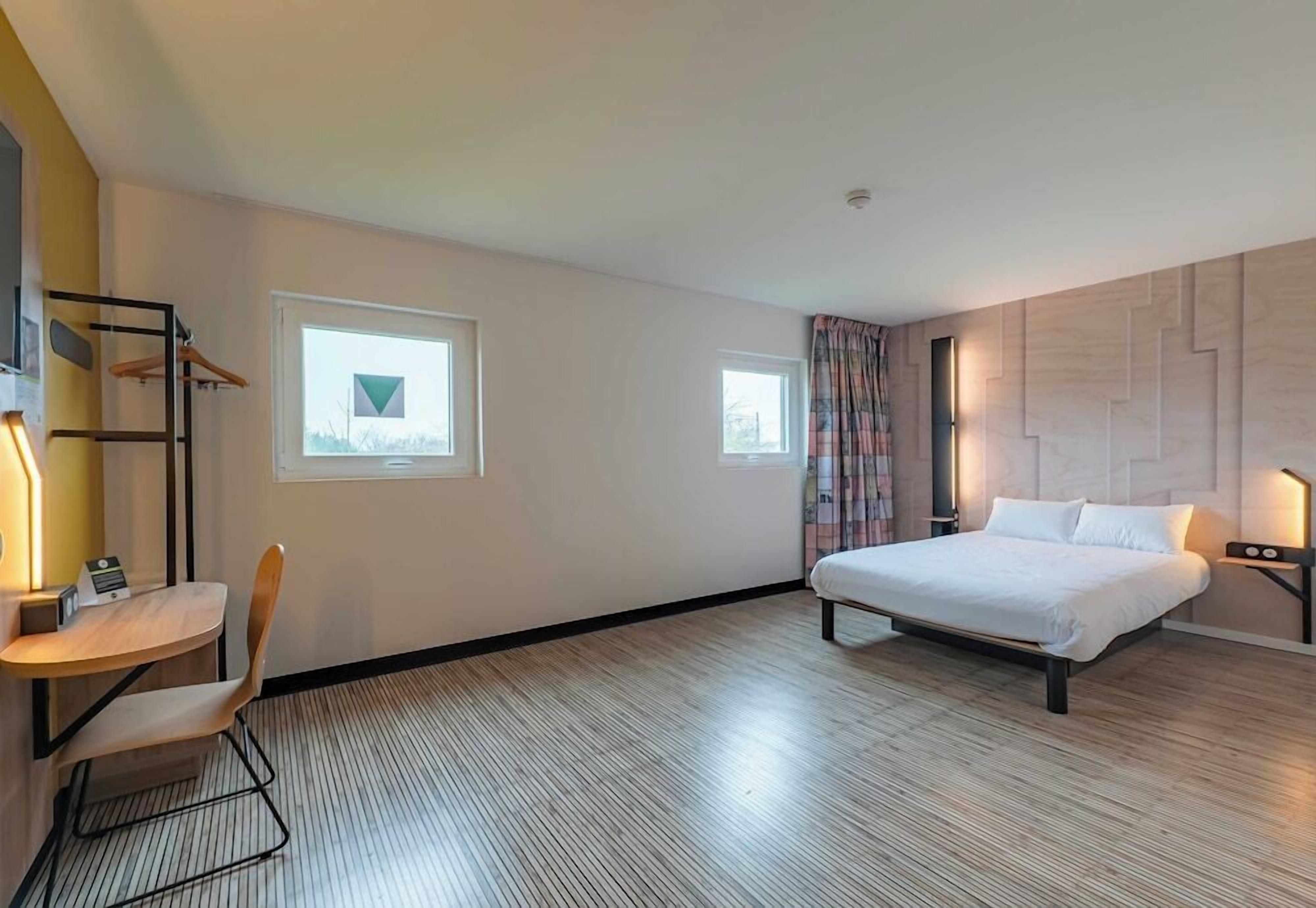 Una habitación de hotel con una cama, un escritorio con una silla, dos ventanas y una lámpara en la pared.