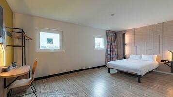 Una habitación de hotel con una cama, un escritorio con una silla, dos ventanas y una lámpara en la pared.