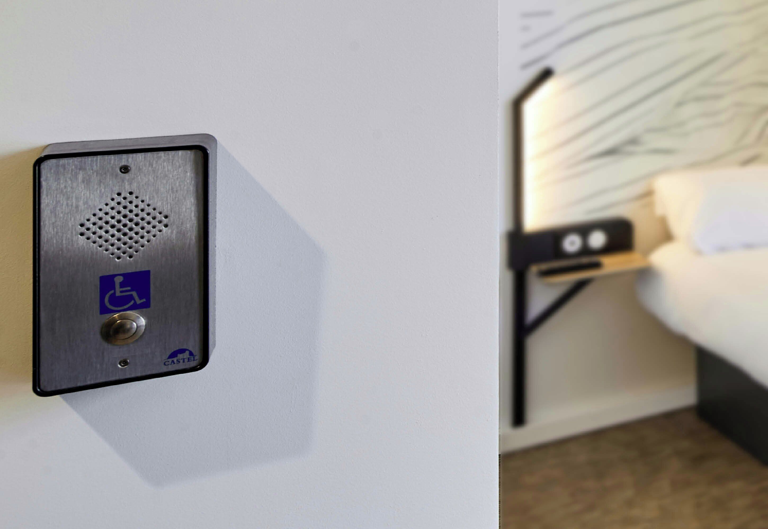 Una habitación de hotel con cama, mesita de noche y un intercomunicador en la pared.