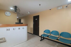 Un área de recepción con un mostrador identificado como 'MUBG', un reloj y una fila de sillas azules.