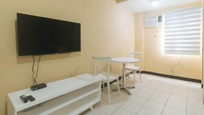 A room with a flat-screen TV mounted on the wall, a white TV stand with remote controls, a small round table with two white chairs, and a window with blinds. - RedDoorz @ MUBG Apartelle near Baclaran Church (Paranaque)