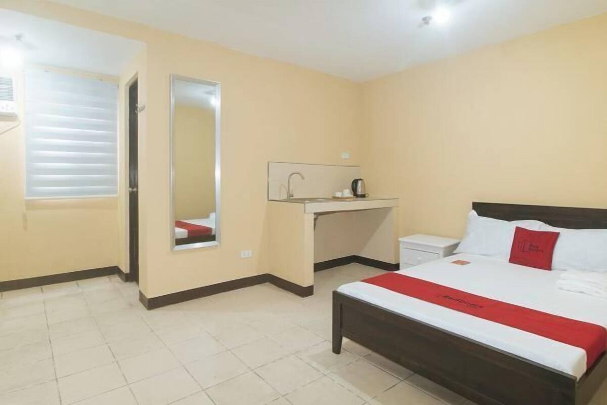 Habitació d'hotel amb un llit, una taula de treball amb un mirall, un lavabo i una finestra amb persianes.