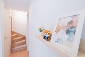 A hallway with a wooden staircase, a small shelf with a teddy bear and a flower, and a framed abstract painting on the wall. - New Garfieldhome Shinjuku (Tokyo)