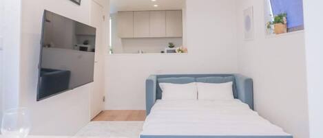 A bedroom with a bed, a flat-screen TV, a small kitchen area, and a window.