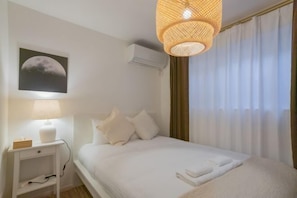 A neatly arranged hotel room with a bed, a bedside lamp, a wall-mounted air conditioner, and a window with curtains.