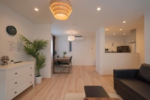 A modern living room with a sofa, a dining area with a table and chairs, a potted plant, and a ceiling light fixture.