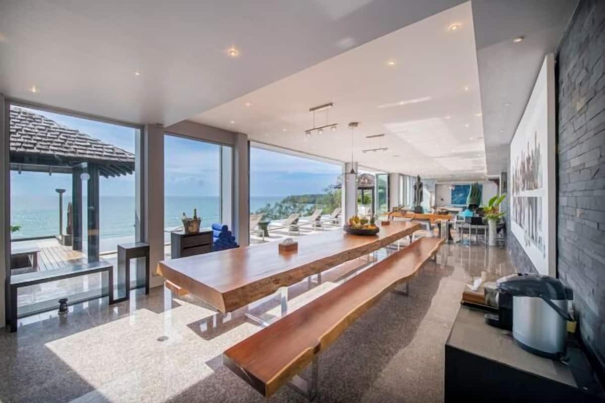 Una cocina amplia con una larga mesa de madera, grandes ventanales que ofrecen vistas al océano y una pared de acento de piedra.
