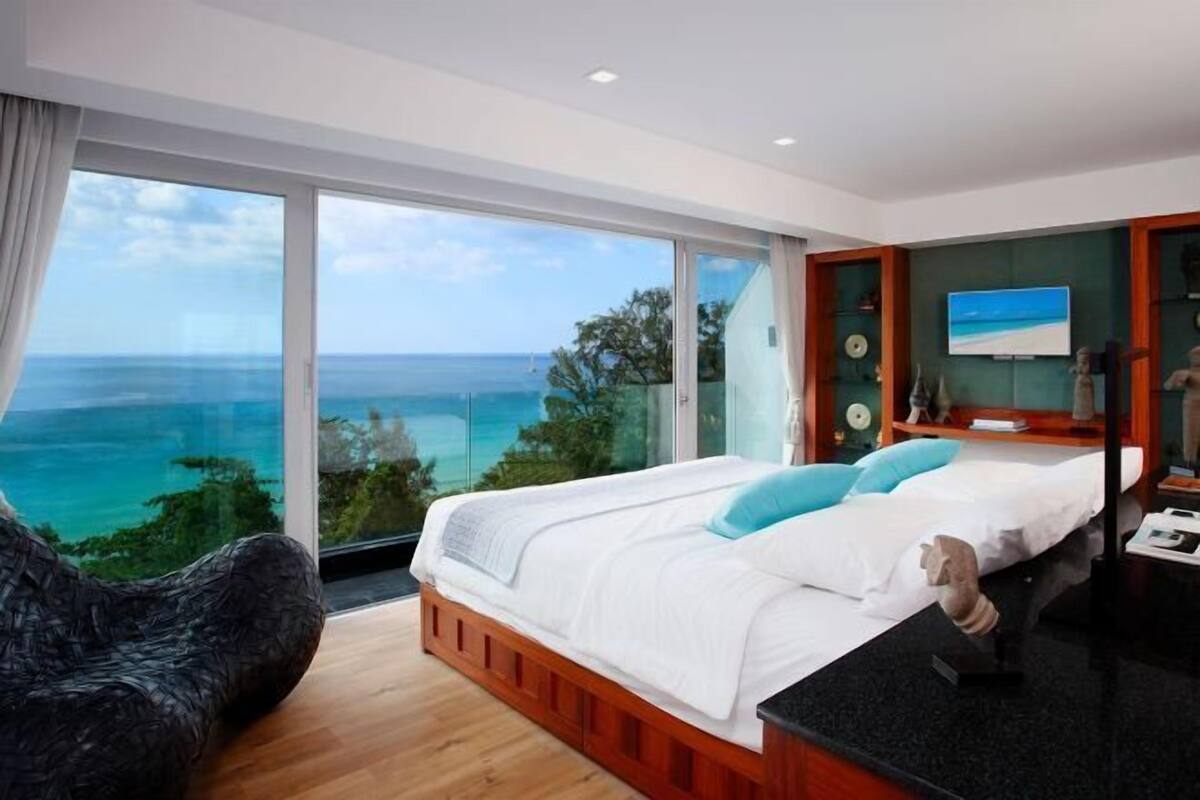 Un dormitorio con un ventanal grande, una cama, un sillón y una mesita con una escultura.