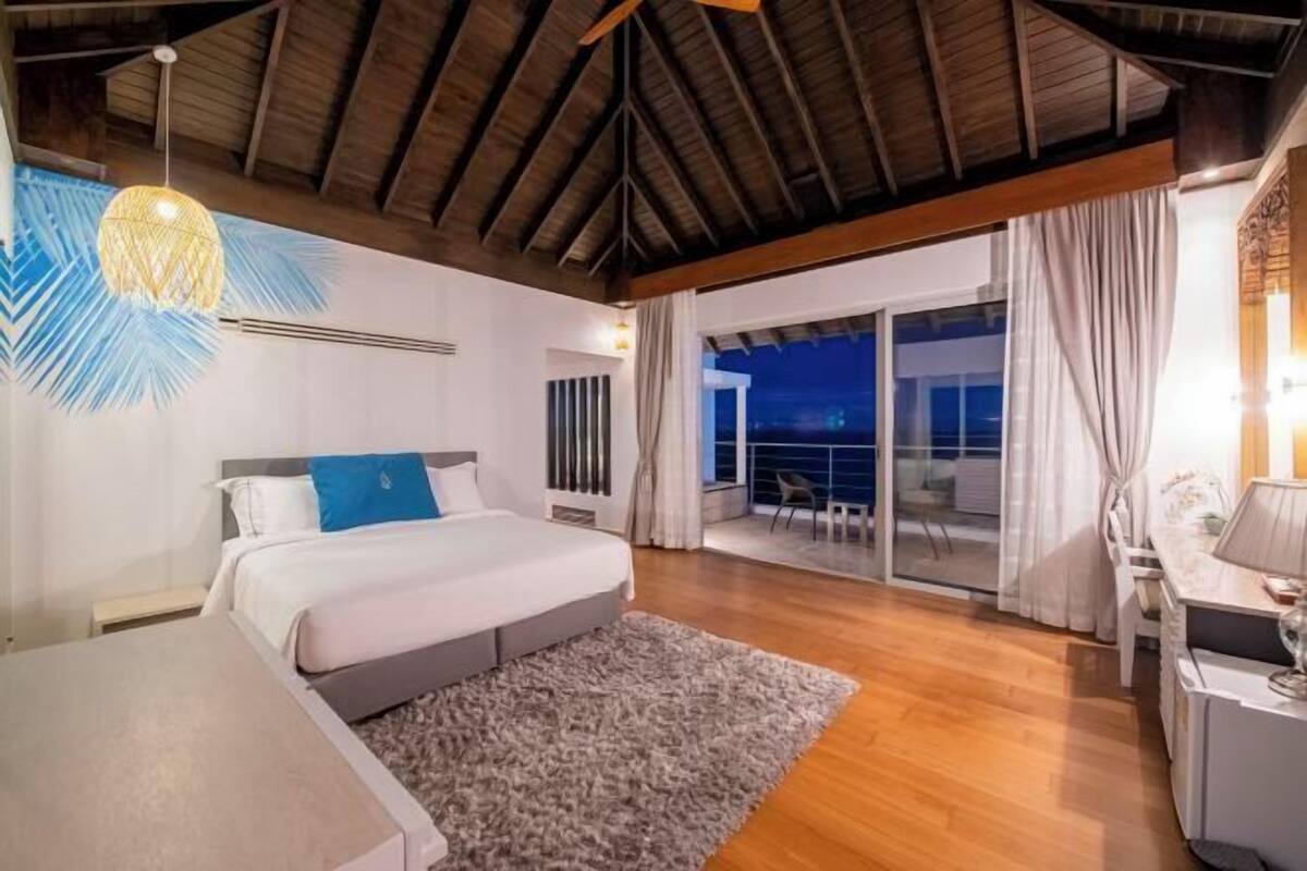Un dormitorio con cama, escritorio, silla, ventana grande y balcón.