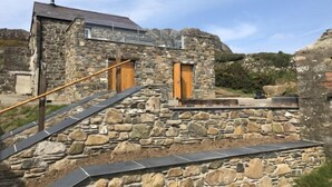 Un bâtiment en pierre avec un escalier menant à une porte en bois, le tout entouré d’un terrain rocheux.
