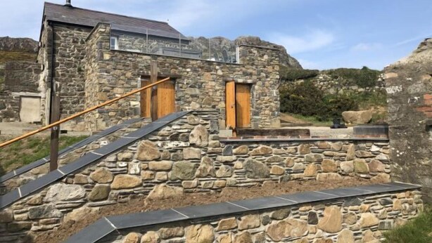 A stone building with a staircase leading to a wooden door, surrounded by rocky terrain. - Tan y Garn - 2 Bedroom Cottage - Whitesands (Saint David's and the Cathedral Close)