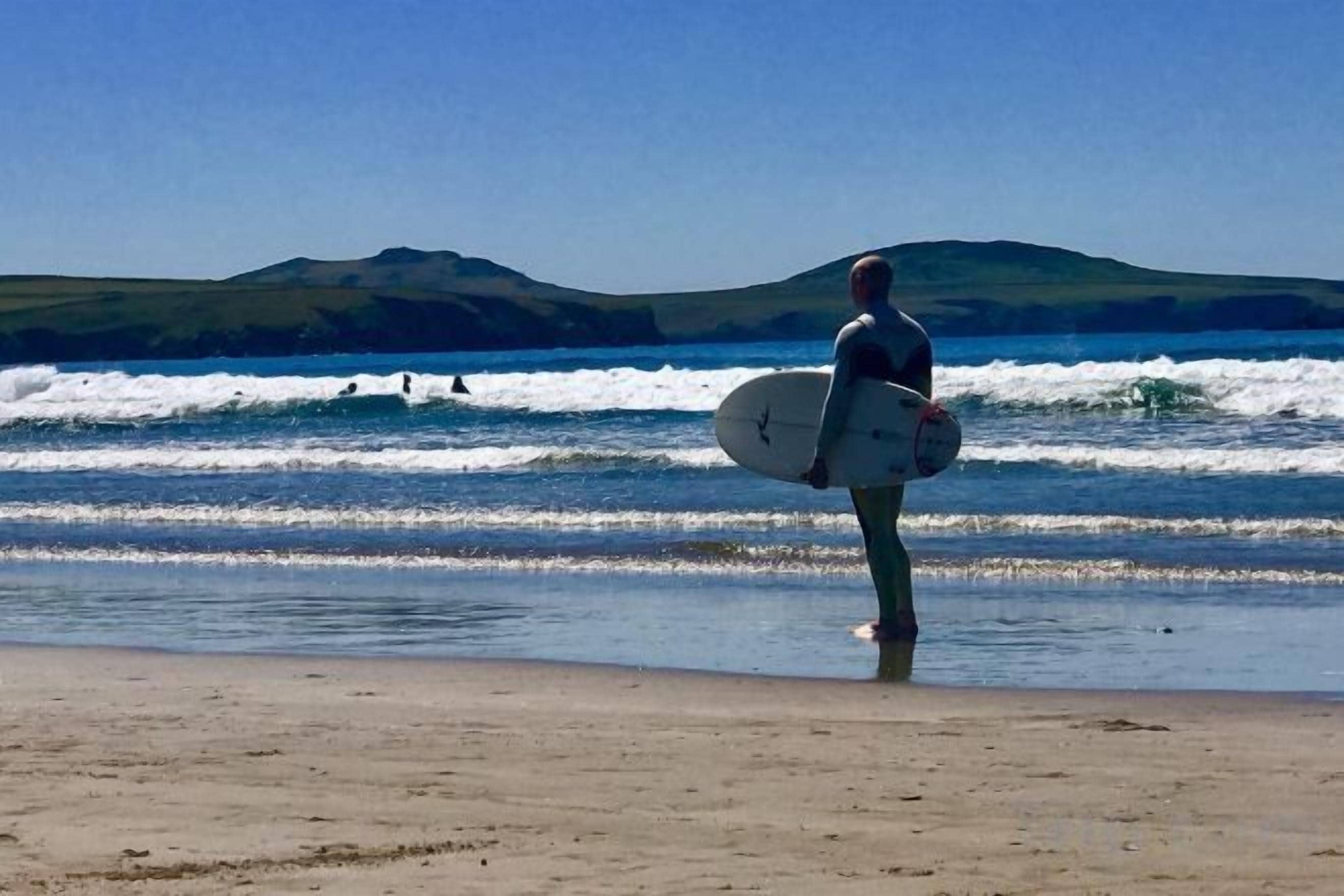 A person carrying a surfboard on a beach with waves and hills in the background.