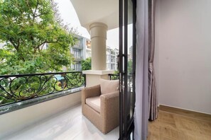Un balcon avec un fauteuil en osier, une porte vitrée et une vue sur la verdure et les bâtiments.