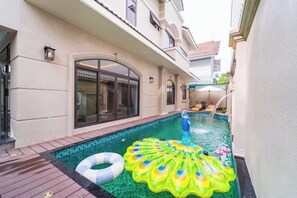 Une piscine avec un flotteur circulaire, une terrasse en bois et un bâtiment doté d’une grande fenêtre et d’une porte.