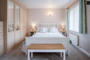 Une chambre à coucher comprenant un lit, des tables de chevet, un banc, une armoire et une fenêtre avec des rideaux.