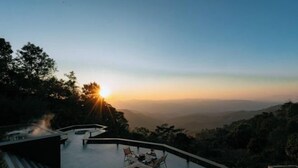 A sunset view from a rooftop infinity pool with a table and chairs, surrounded by trees and hills. - Misty Valley Doichang (Mae Suai)