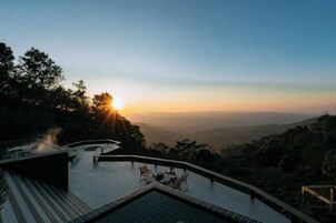Une vue sur le coucher de soleil depuis une piscine à débordement située sur un toit, avec une table et des chaises, le tout entouré d’arbres et de collines.