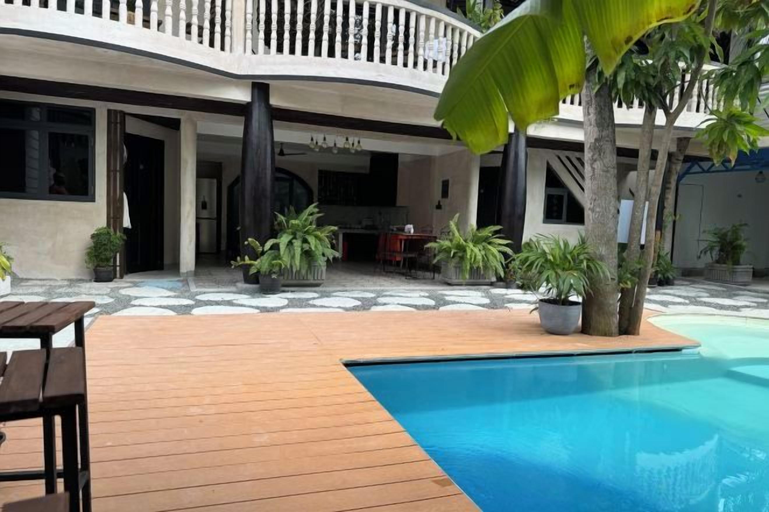 Zona de piscina con cubierta de madera, plantas en macetas y comedor con mobiliario exterior.