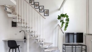 Ein modernes Interieur mit einer weißen Wendeltreppe, einem Schreibtisch mit Stuhl, einer hängenden Pflanze und einem Fernseher auf einem Podest.