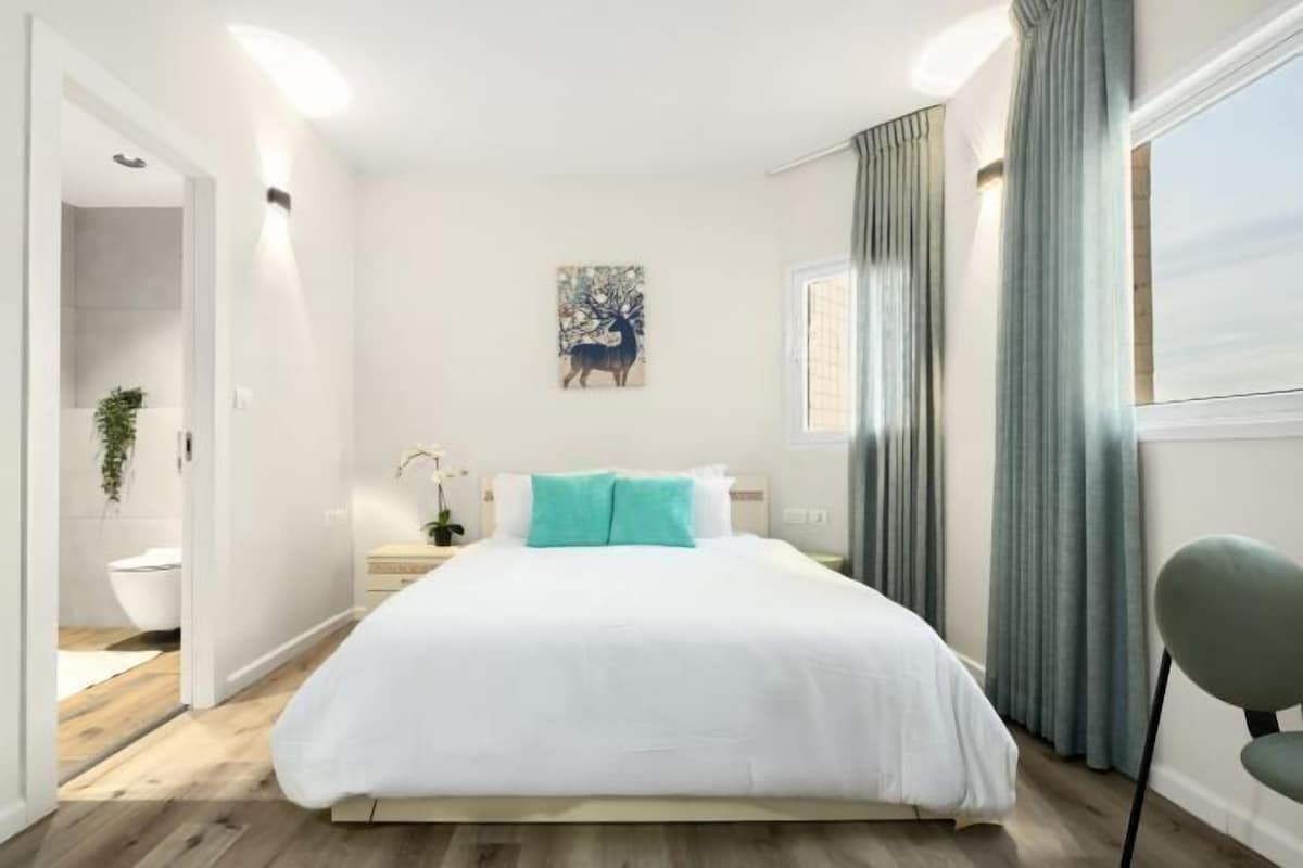 A bedroom with a bed, a nightstand, a wall-mounted artwork, and a window with curtains.