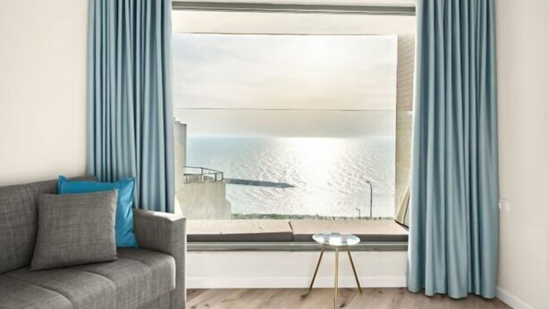 A modern living room with a grey sofa, a small round table, and a view of a body of water through a large window. - Netanya Beach & Pool Elegant Stay (Netanya)
