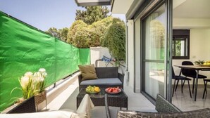 A balcony with a sofa, dining table, and chairs, a green screen, and a glass door leading to a room. - Bahai Gardens Serenity Apartment (Haifa)