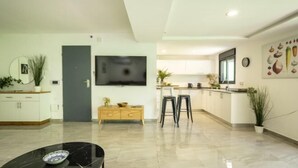 A modern living room with a flat-screen TV, a coffee table, and a dining area with bar stools. - Bahai Gardens Serenity Apartment (Haifa)